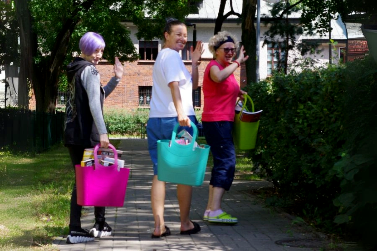 Trzy osoby na świeżym powietrzu, każda z nich niesie kolorową torbę wypełnioną książkami i radośnie macha ręką w otoczeniu parku.