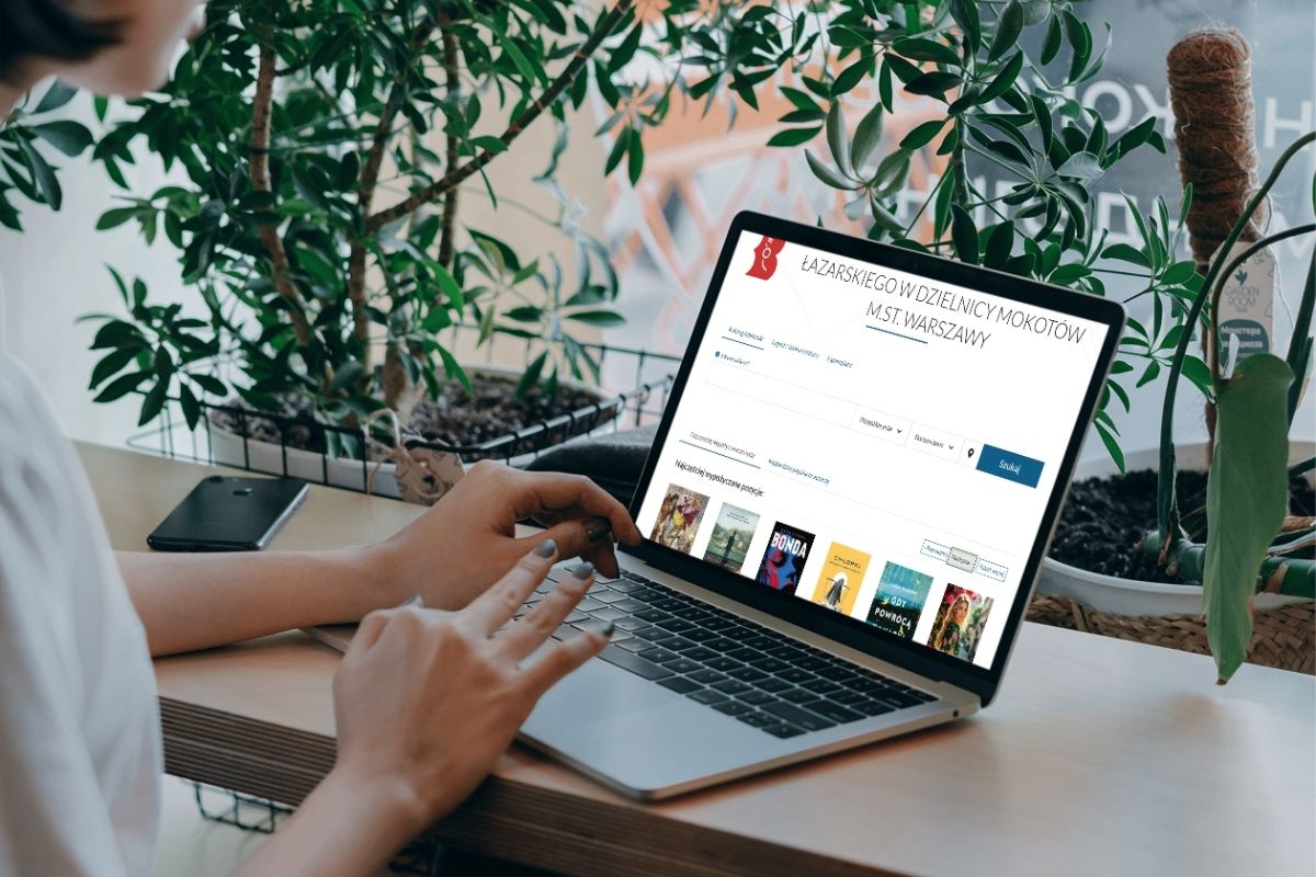 Kobieta siedzi przy oknie pełnym kwiatów i przegląda katalog biblioteki w komputerze