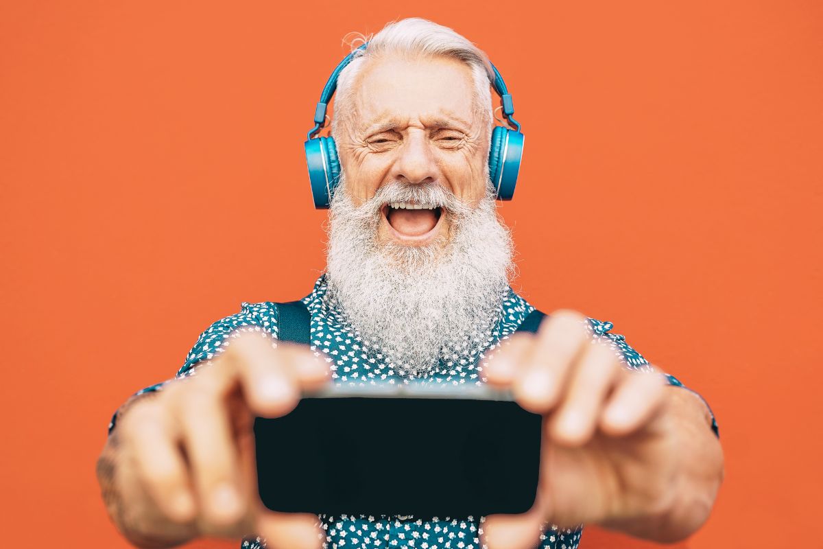 Na pomarańczowym tle mężczyzna z brodą i w słuchawkach na uszach robi sobie selfie smartfonem