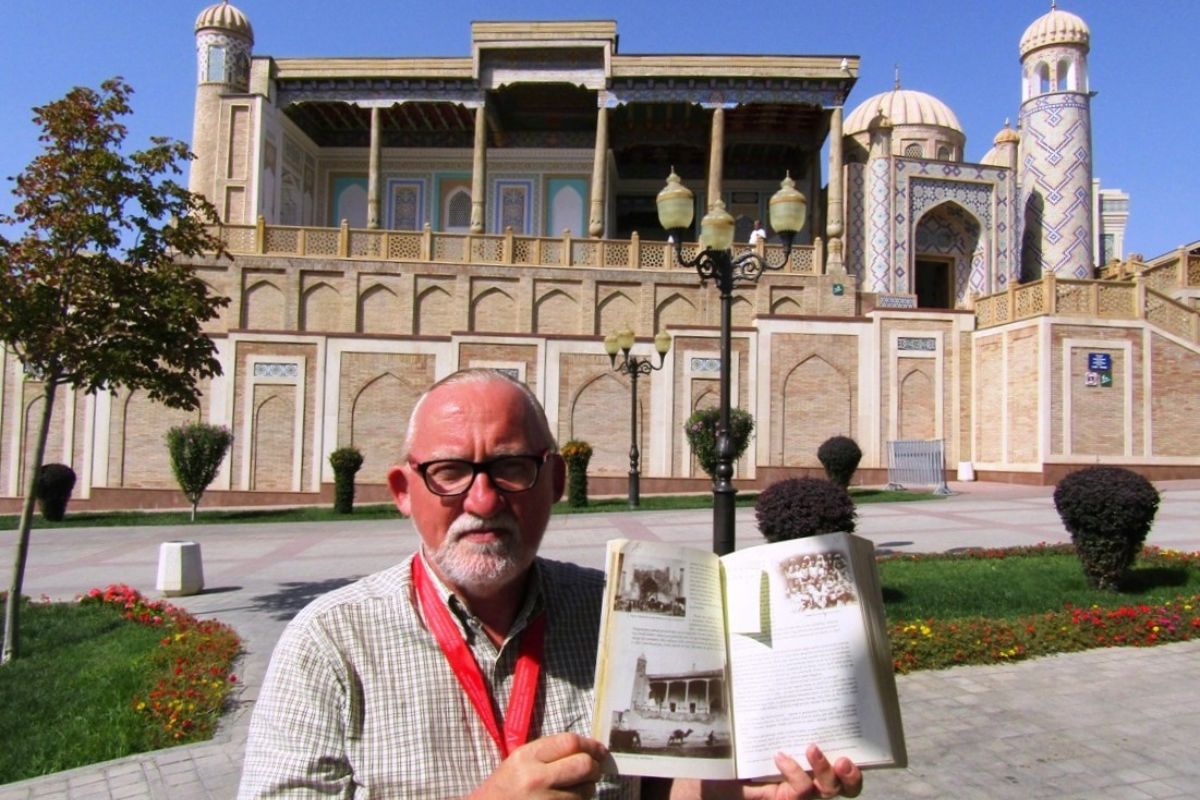 Mężczyzna w okularach prezentuje książkę, w tle egzotyczna budowla