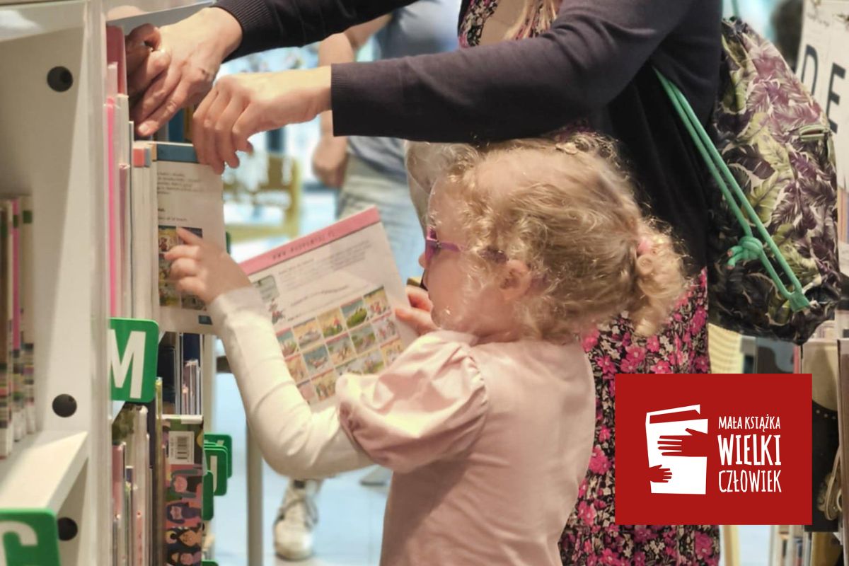 Dziewczynka z jasnymi włosami wkłada książkę na półkę w bibliotece, w tle jej mama